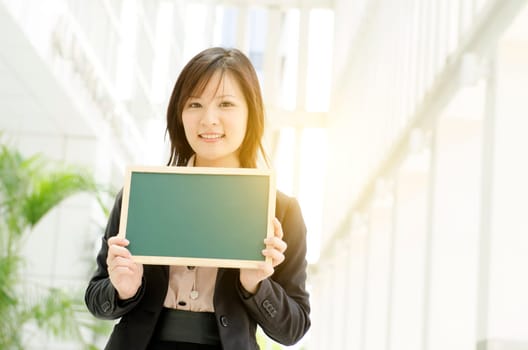 Portrait of a young Asian business woman smiling and holding a blank green chalkboard, standing at an office environment, beautiful golden sunlight at background.