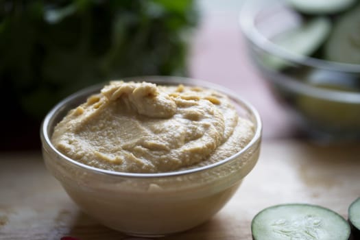 Bowl of hummus on table with vegetables out of focus in background.