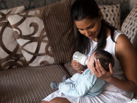 Young mother sitting on her sofa whilst feeding her baby son a bottle.