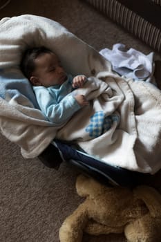 cute, baby boy is fast asleep in his car seat. He is wrapped up in a fluffy blanket with a teddy beside him.