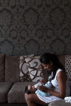 Young mother is sitting on her sofa with her baby son lying in her lap. He is looking at the camera whilst his mother feeds him a bottle.