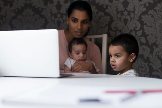 Young mother keeping her children entertained by showing them something on her laptop. She is holding her baby son and the eldest is standing next to her, looking at the screen.
