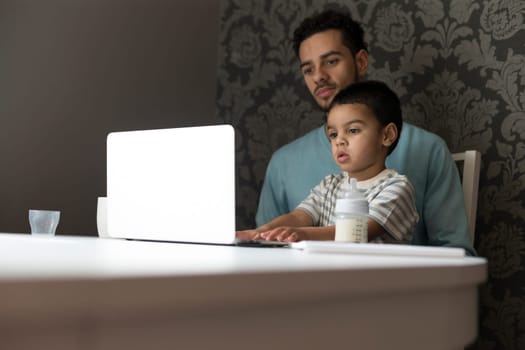 Young father is sitting at the table with his son on his lap. His son is copying his dad and exploring his curiosity by pressing buttons and looking at the screen.