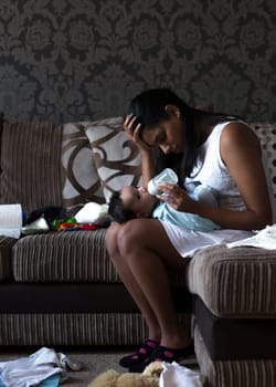 Stressed young mother sitting on her sofa whilst feeding her baby son. She has her head in her hand and is surrounded by mess.