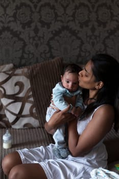 Young mother is sitting on her sofa and holding her baby son who has just been fed. She is kissing him on the head with him held close to her.