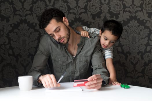 Little boy is climbing on his dad, playing with his toy car. His dad is trying to make bill payments through the phone.