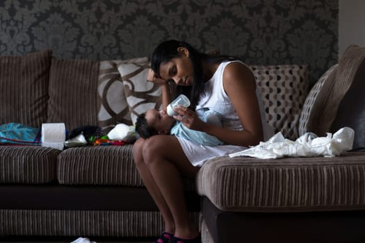 Stressed young mother sitting on her sofa whilst feeding her baby son. She has her head in her hand and is surrounded by mess.