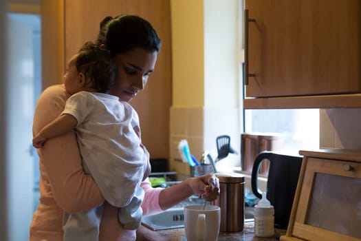 Young mother making a cup of tea with her baby son on her shoulder