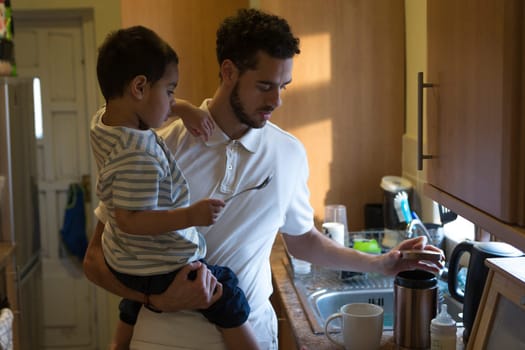 Young father making a cup of tea with the help of his son. He is opening a pot of sugar and the little boy is holding the spoon.