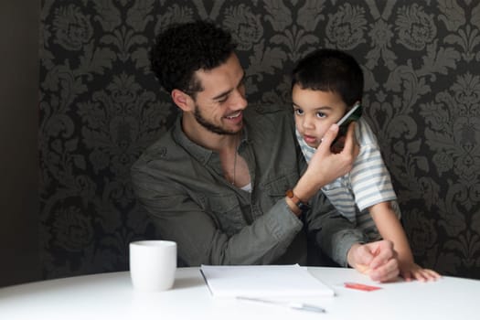 Young father is laughing as his son copies him. He is holding the phone to his sons ear, who is pretending to talk to someone.