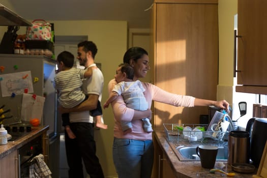 Young family in the kitchen. The mother is washing dishes with their baby on her shoulder while the father is looking in the fridge with their eldest on his shoulder.