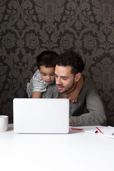 Little boy is climbing on his dad, trying to help him with his internet banking. They are sitting behind a laptop at home.