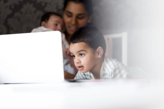 Little boy watching cartoons on his mothers laptop while she feeds her baby son in the background