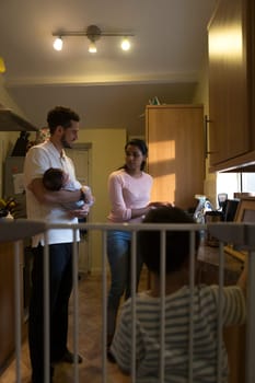 Young family in the kitchen. The mother is washing dishes and talking to her partner, who is holding their baby son, The eldest son is standing with his back to the camera, playing with his toy car on the worktop.