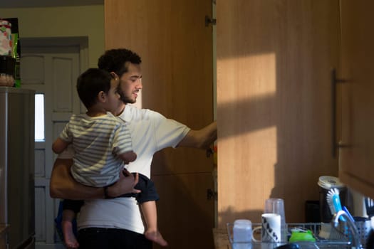 Young father in the kitchen, getting something out of the cupboard with one hand with his son on his shoulder.