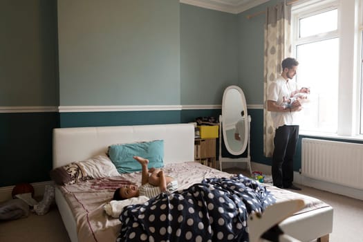 Little boy rolling around on his father's bed, who is standing by the window holding his upset baby son