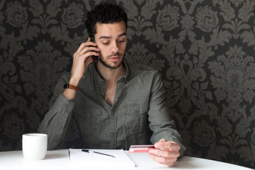 Young man sitting at the table at home, using his smartphone and his bank card to pay bills over the phone.