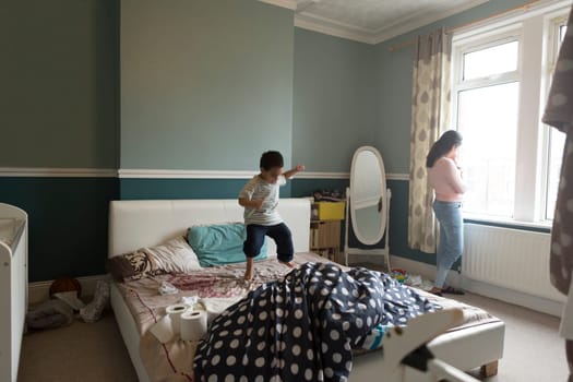 Little boy is jumping on his mother's bed, who is standing by the window with her baby son in her arms.