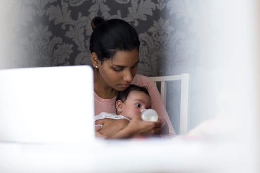 Young mother feeding her baby son a bottle of milk whilst sitting at her computer.