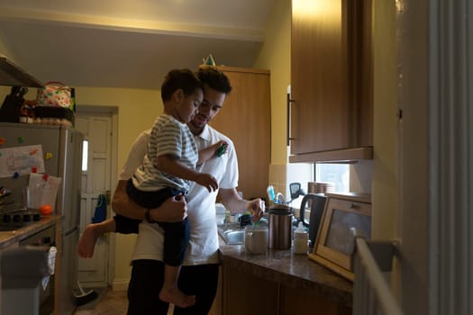 Young father making a cup of tea with his son on his shoulder