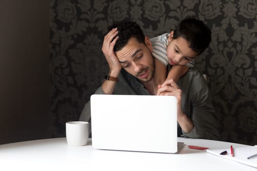 Little boy is climbing on his dad, trying to help him with his internet banking. They are sitting behind a laptop at home.