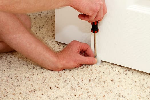 Caucasian hands of an adult male using a screwdriver to secure a white plastic closet door guide into a carpeted floor of a home. Securing a closet door floor guide