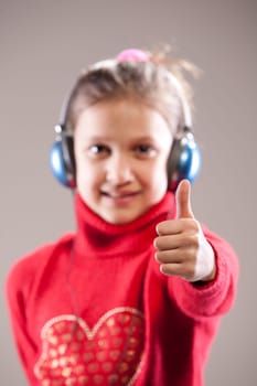 Little girl listening to the music inher blue headphones and showing her thumb up (IN FOCUS) the face is out of focus