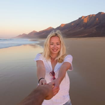 Young romantic couple, holding hands, having fun on perfect deserted beach at sunset. Shot from boyfrieds perspective. Guy looking at her beautiful carefree girlfriend.
