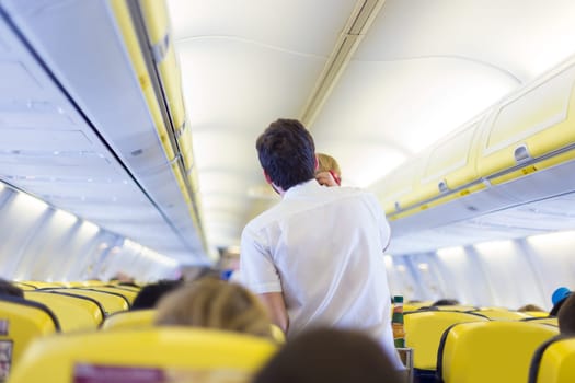 Interior of airplane with passengers on seats and steward walking the aisle.