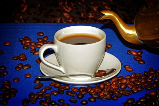 White cup of coffee on blue tablecloth with coffee beans and neck of a golden jug