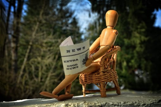 Brown wooden figure sits on terrace in rattan armchair and reads newspaper,fuzzy nature background
