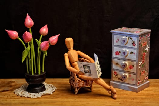 Wooden figure reading newspaper in the living room, next to dresser and wooden flowers with dark background