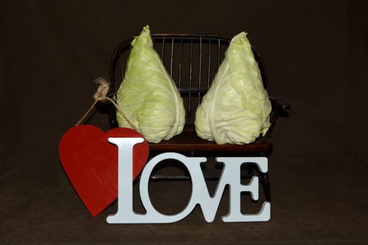 Two pointed cabbage heads sit on wooden bench against a dark background with red heart and white love lettering in front of it