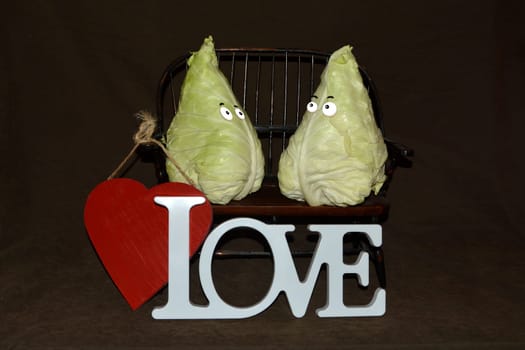 Two pointed cabbage heads sit on wooden bench against a dark background with red heart and white love lettering in front of it