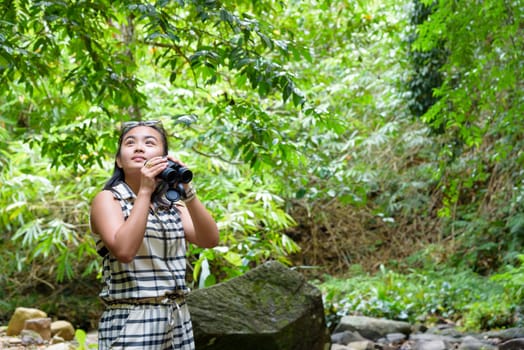 Beautiful young girl hiking is using binoculars look for birds in tropical forest near the waterfalls in Thailand