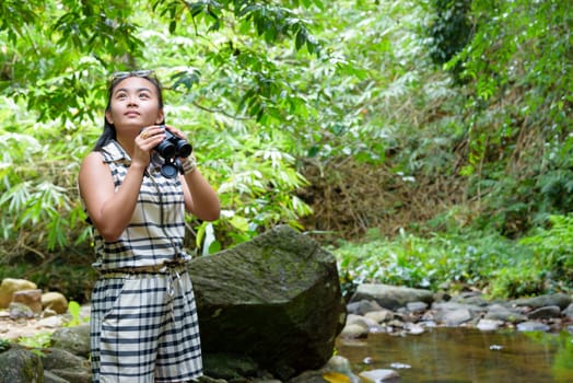 Beautiful young girl hiking is using binoculars look for birds in tropical forest near the waterfalls in Thailand