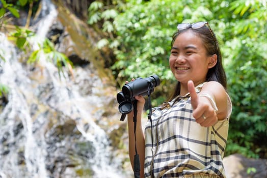 Beautiful young girl hiking is using binoculars look for birds smiled and raised a thumb to the camera in tropical forest near the waterfalls in Thailand