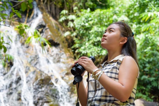 Beautiful young girl hiking is using binoculars look for birds in tropical forest near the waterfalls in Thailand