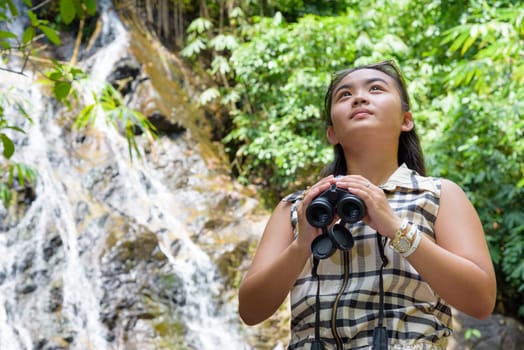 Beautiful young girl hiking is using binoculars look for birds in tropical forest near the waterfalls in Thailand