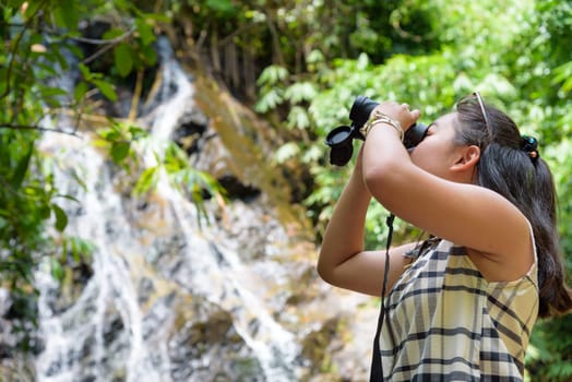 Beautiful young girl hiking is using binoculars look for birds in tropical forest near the waterfalls in Thailand