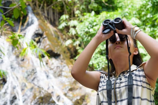 Beautiful young girl hiking is using binoculars look for birds in tropical forest near the waterfalls in Thailand