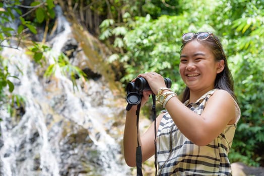 Beautiful young girl hiking is using binoculars look for birds smiling to the camera in tropical forest near the waterfalls in Thailand