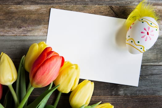 Colorful red and yellow tulips and Easter egg, with a blank card on rustic wooden table.