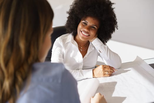 Team of two female architects, sitting at desk in office. The women talk and smile reviewing a building plan. Medium shot
