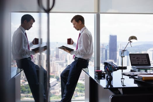 Adult businessman sitting on desk in modern office and reading news on tablet pc with a cup of coffee. The man looks out of the window and contemplates the city and skyscrapers.