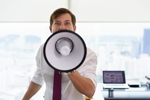 Portrait of furious business man screaming with megaphone in modern office. The angry man looks at camera. Copy space
