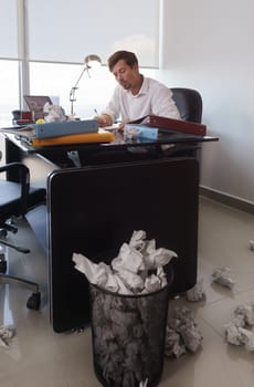 Corporate manager in modern office tries to write a job letter. The man is frustrated and keeps on throwing paper on desk and trash bin