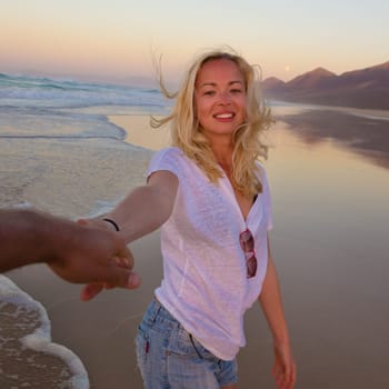 Young romantic couple, holding hands, having fun on perfect deserted beach at sunset. Shot from boyfrieds perspective. Guy looking at her beautiful carefree girlfriend.