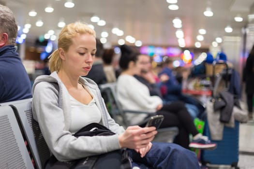 Casual blond young woman using her cell phone while waiting to board a plane at the departure gates. Wireless network hotspot enabling people to access internet conection. Public transport.