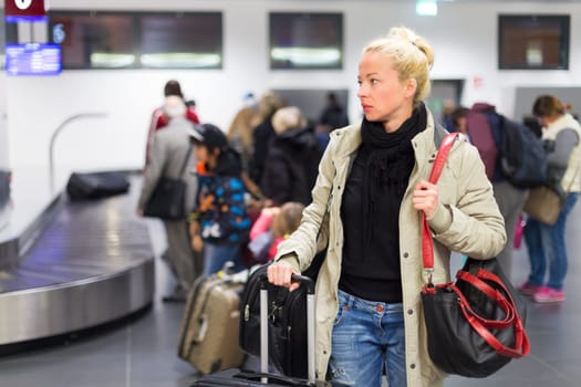 Casually dressed young female traveller waiting in airport arrival terminal hall,  collecting her laggage . Blured background. Can also be used as railway, metro, bus station.